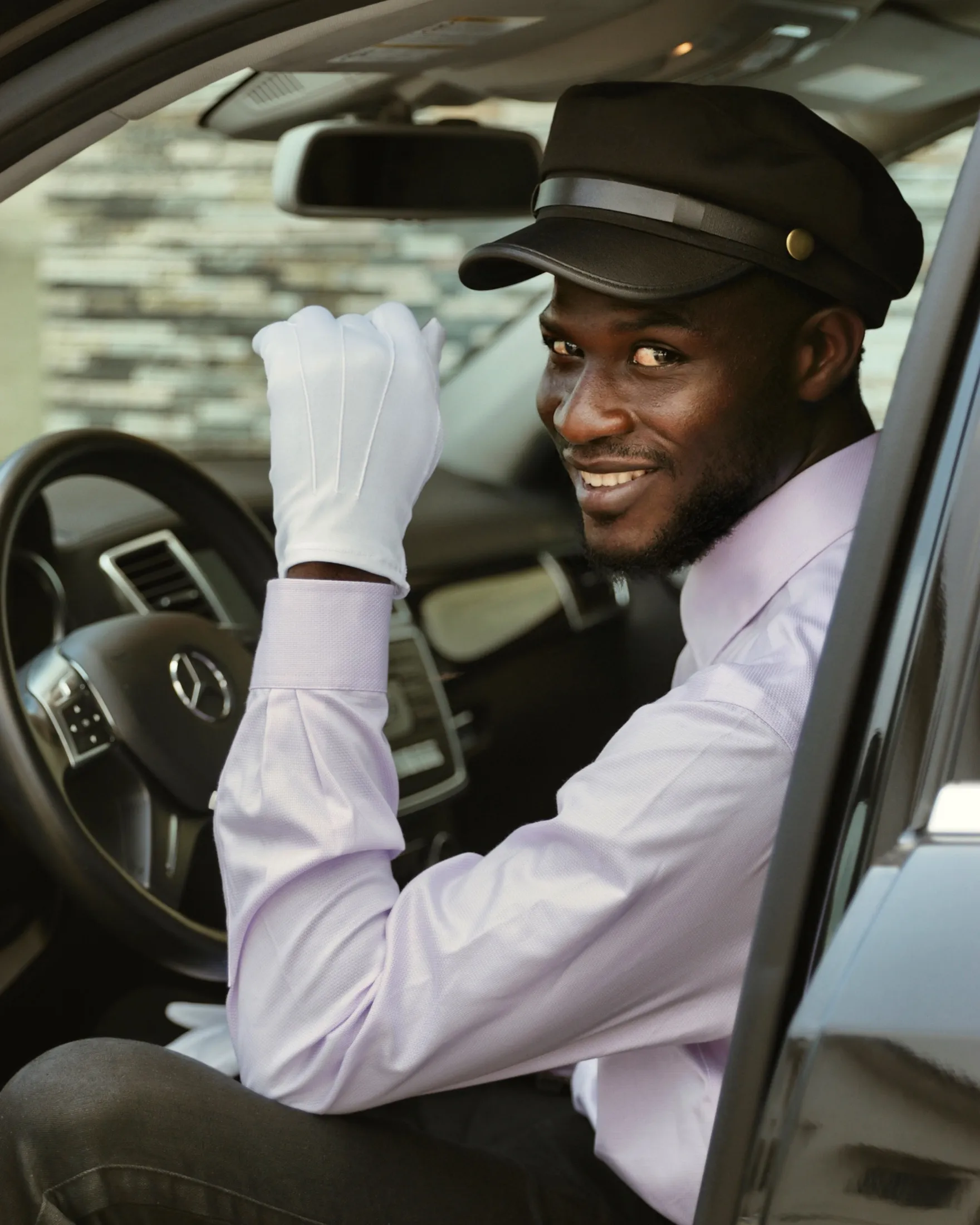 Chauffeur in a cap and white gloves smiling from the driver’s seat of a black car.