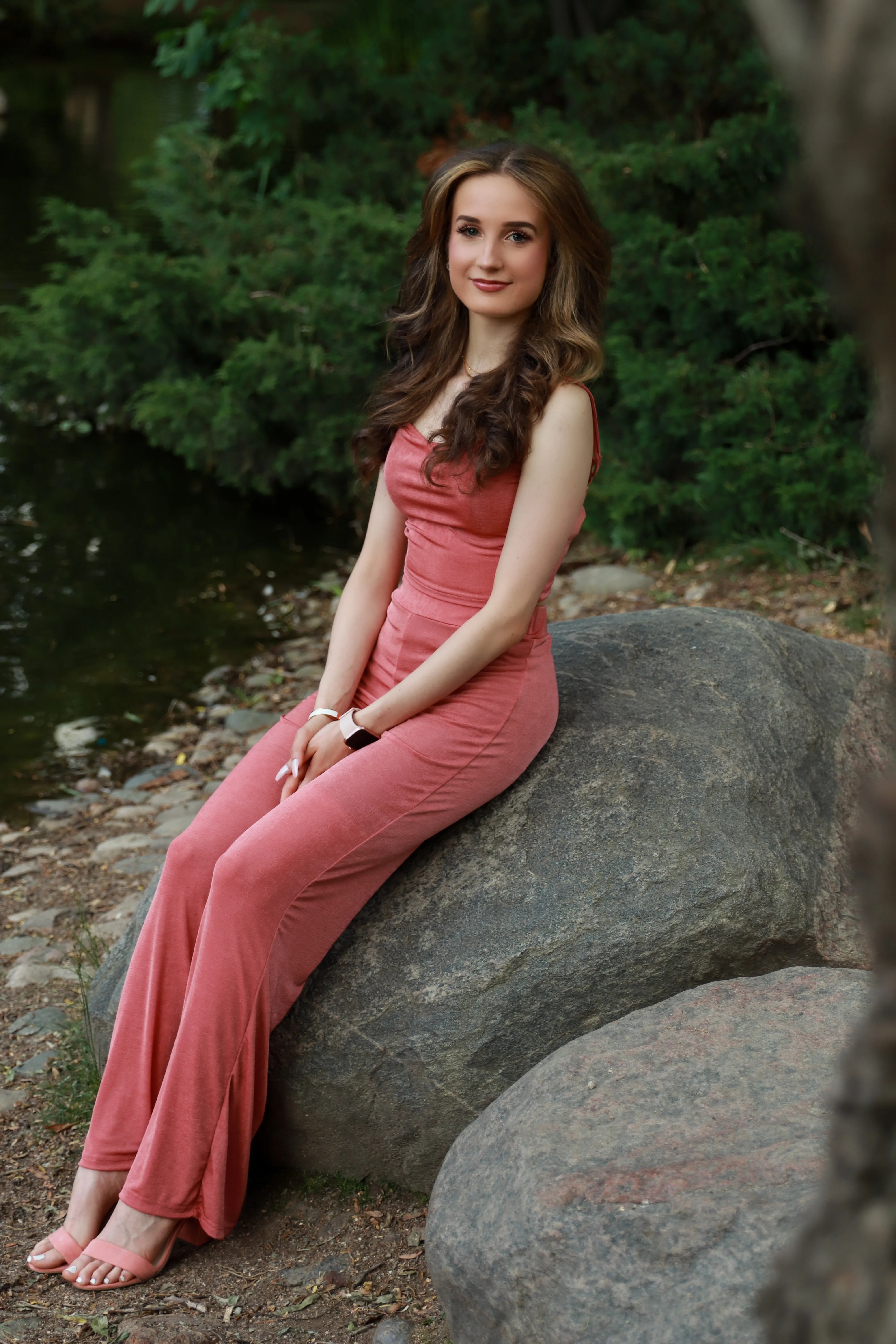 Woman in a rose-pink jumpsuit seated on a large rock beside a pond.